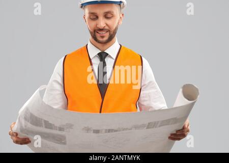 Raccolto di giovane architetto bearded in arancione riflettente giubbotto e casco tenendo grande mappa. Ingegnere sorridente che analizza il piano di costruzione, isolato su grigio. Concetto di architettura, costruzione. Foto Stock