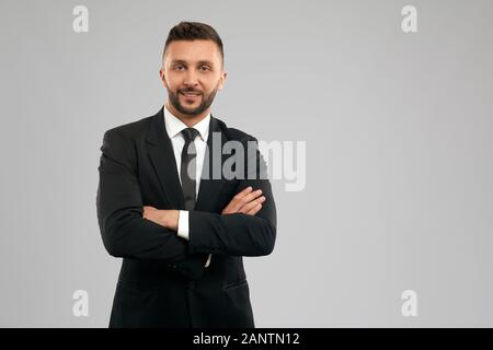 Vista frontale del giovane uomo d'affari sorridente in abito nero formale che posa con le mani incrociate. Lavoratore bruna di successo in piedi e guardando la macchina fotografica, isolato su sfondo grigio. Concetto di business. Foto Stock