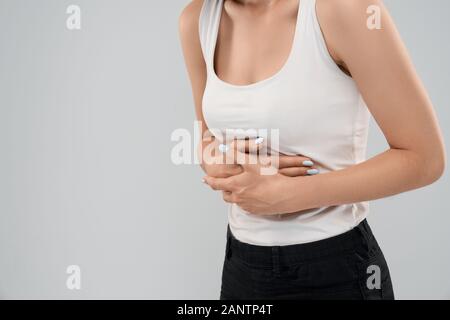 Clolse fino di piegatura in incognito caucasian donna in abito casual in posa su grigio Sfondo isolato e toccando parte superiore dello stomaco a causa di ache. Parte del raccolto di bruna che soffrono di dolore addominale. Foto Stock