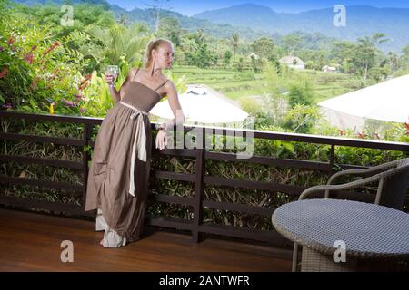 Giovane bella donna in una lunga sundress beige con un bicchiere di vino si affaccia sulla sua spalla dalla terrazza per la natura tropicale Foto Stock