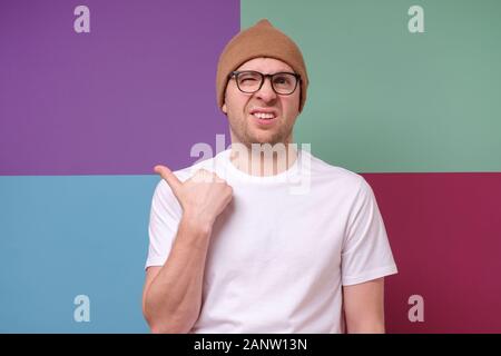 Sospette uomo millenario che indossano occhiali punti il dito oltre al discutibile diffidenti annuncio, copia vuota spazio su sfondo colorato. Studio Foto Stock