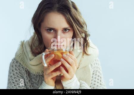 Ritratto di mal donna moderna in rotolo maglione collo e cardigan bicchiere di tè caldo con lo zenzero, succo di limone e il miele sulla luce invernale sfondo blu. Foto Stock