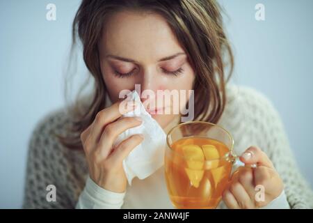 Ill elegante massaia in rotolo maglione collo e cardigan bicchiere di tè caldo con lo zenzero, succo di limone e il miele contro luce invernale sfondo blu. Foto Stock