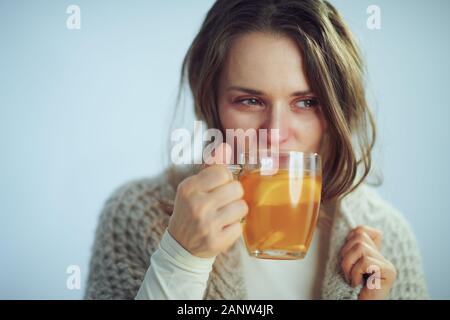 Ill elegante massaia in rotolo maglione collo e cardigan bicchiere di tè caldo con lo zenzero, succo di limone e il miele sulla luce invernale sfondo blu. Foto Stock