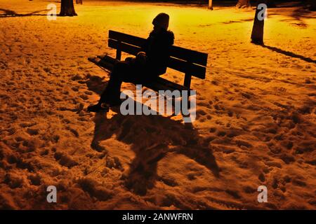 Silhouette di una signora su una panchina del parco bagnata dal bagliore arancione di una lampada da strada nel parco locale, in una serata invernale e innevata. Ottawa, Ontario, Canada. Foto Stock