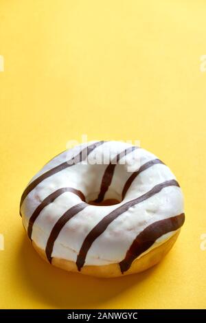 Una ciambella rotonda singola con vetri di ghiaccio bianco e strisce di cioccolato marrone scuro su uno sfondo giallo con spazio di copia. Immagine verticale Foto Stock