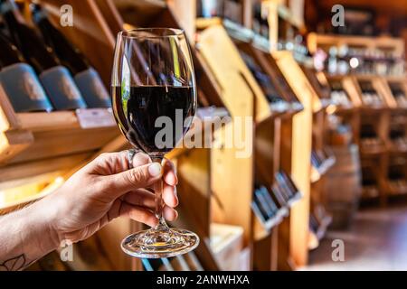 Mano che tiene un bicchiere di vino rosso fuoco selettivo vista, sala degustazione vini bottiglie visualizzazione su scaffalature in legno Scaffali sfondo, wine shop interno Foto Stock