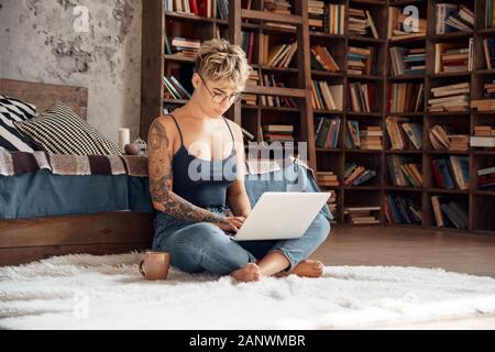 Lato Hustle. Giovane donna capelli corti in bicchieri seduto sul piano di lavoro on-line su laptop concentrato con tazza di caffè Foto Stock