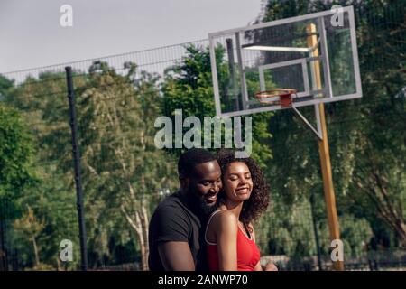 Attività all'aperto. Coppia africana in piedi sul campo da pallacanestro avvolgente gioioso sorridente Foto Stock