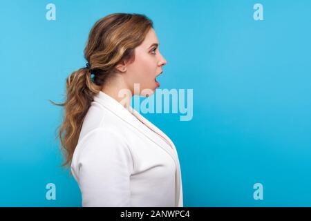 Wow, incredibile! Vista laterale di stupiti donna con la coda di cavallo pettinatura in camicia bianca cercando con la bocca aperta a sorpresa, sconvolta da crazy news. interna s Foto Stock