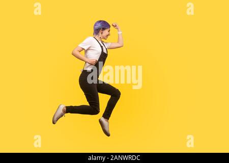 Vista laterale a lunghezza piena, entusiasta tanga energica ragazza con i capelli viola in tuta a saltare o a volare in aria, esegue rapidamente veloce, fretta per s Foto Stock