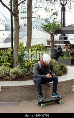 Giovane uomo con skateboard elettrico potenziato Mini X e controllo bluetooth intorno al polso seduto e fare una pausa nella South Bank Londra, Regno Unito Foto Stock