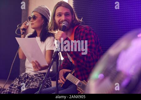 Ritratto di con i capelli lunghi giovane a suonare la chitarra e cantare al microfono durante le prove o di concerto con la banda musicale in ambienti con scarsa illuminazione studio, spazio di copia Foto Stock