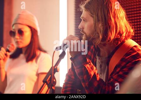 Vista laterale ritratto di con i capelli lunghi giovane a suonare la chitarra e cantare al microfono durante le prove o di concerto con la banda musicale in ambienti con scarsa illuminazione studio, spazio di copia Foto Stock