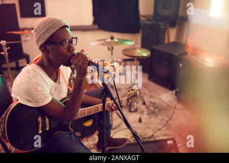 Angolo alto ritratto di contemporaneo afro-uomo cantano al microfono e suonare la chitarra durante le prove o di concerto con la banda musicale in studio di registrazione, spazio copia Foto Stock