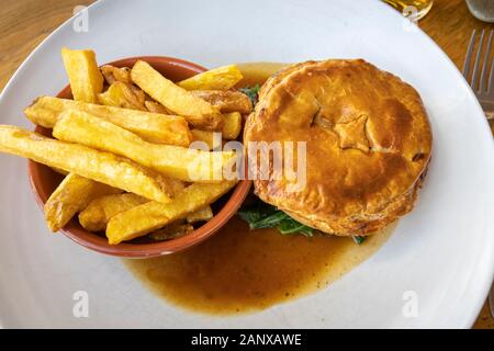 Tipica colazione nutriente e gustosa British pub grub pasto: caldo Torta a base di carne in salsa servita con una ciotola di golden fried chunky chip su un bianco piastra di cina Foto Stock