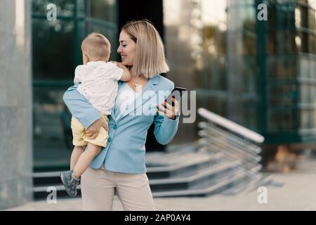 Donna d'affari di successo in tuta blu con il bambino. Madre di successo in affari a piedi con il bambino nelle sue mani e nel suo messaggio sms sul cellulare Foto Stock