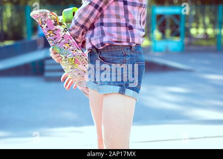 Ritratto di una bambina, in un plaid shirt, in piedi con uno skateboard, nel parco giochi. Teen ragazza sorridente. I bambini attivi lifestyle Foto Stock