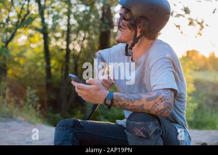 Sorridenti ciclista uomo utilizzando i moderni smartphone al di fuori in appoggio dopo intensi in sella a una moto, sportivo attiva il controllo dei suoi risultati fitness via cel Foto Stock