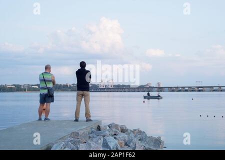 Due persone sono in piedi sul molo, vicino al fiume. Guardare i pescatori pescare da una barca nel fiume. Foto Stock