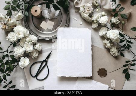 Moody tabella di nozze mockup scena. Composizione di cancelleria con fading white rose fiori, piastra di argento, nero forbici, buste e saluto vuoto ca Foto Stock