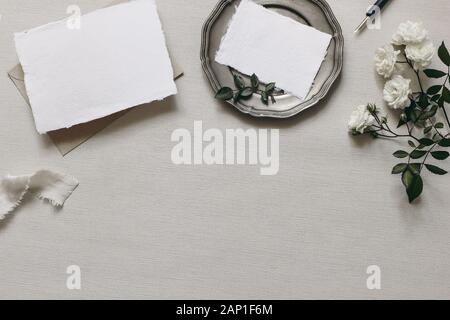 Articoli di cancelleria tabella di nozze mockup scena. Moody composizione con fading white rose fiori, piastra di argento, calligrafia pen, biglietti vuoti e copia Foto Stock