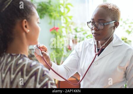 Un nero ragazza medico esamina un paziente messicano in un ufficio medici esegue auscultazione dei polmoni con uno stetoscopio Foto Stock