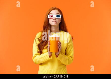 Lo stile di vita, hobby e concetto di persone. Carino geeky redhead donna in 3d carta bicchieri, azienda di popcorn e mordere il labbro come fissando attore preferito giocare Foto Stock