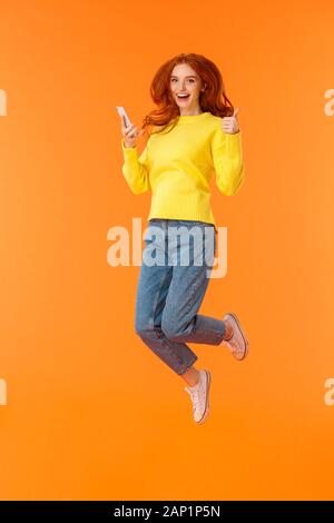 Verticale a tutta lunghezza shot spensierato, lieti di bella ragazza di zenzero in maglione di lana, jeans, saltando su sfondo arancione, tenendo lo smartphone Foto Stock