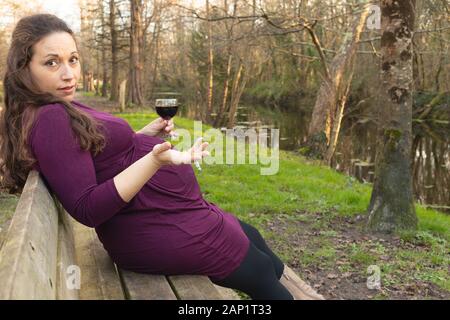 Donna incinta con alcool e sigarette nelle sue mani, seduti su una panchina nel parco e guardando la telecamera con un interrogatorio look e scrollare gesto Foto Stock