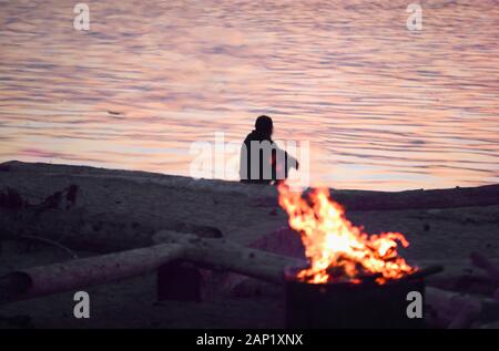 un uomo solitario siede da un falò su una spiaggia meditando e contemplando i suoi pensieri e fissando l'oceano Foto Stock