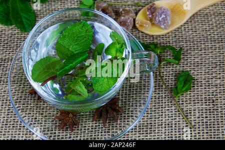 Close up di isolato di tè trasparente in vetro con tazza di foglie di menta fresca, il cucchiaio di legno marrone con caramelle di zucchero rock e anice stelle. iuta sfondo mat. Foto Stock