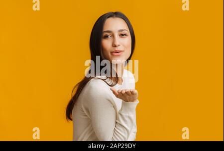 Bella bruna ragazza bacio di soffiaggio in telecamera, ponendo su sfondo giallo Foto Stock