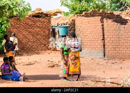 Una donna malawia sta parlando con un amico mentre sostiene un secchio d'acqua sulla sua testa in una posizione di villaggio Foto Stock