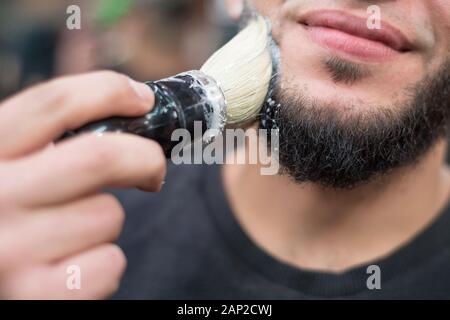 Primo piano di un barbiere di mettere qualche crema di rasatura su un cliente del viso prima di dargli una rasatura la rasatura una barba di un uomo caucasico. Lathers una barba, prepa Foto Stock