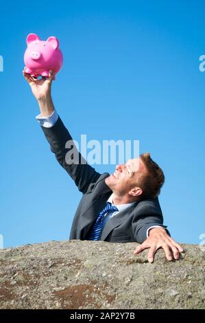 Imprenditore sorridente tenendo premuto su un fido rosa salvadanaio rock come egli raggiunge la cima di una scogliera rocciosa Foto Stock