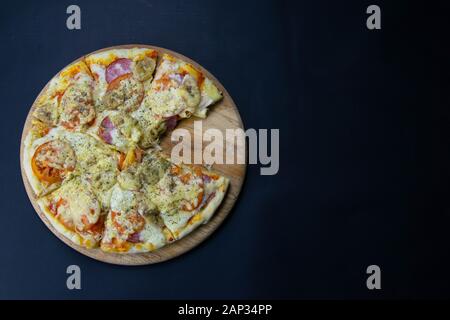 Pizza con salame, mozzarella, pomodori e banane sorge su una tavola di legno, su sfondo nero. Viste dall'alto strega liberare spazio Foto Stock