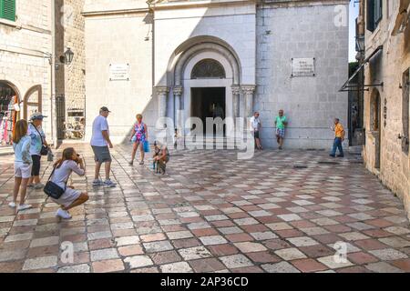 Una donna che mendica per assistenza sui gradini della chiesa come turisti si riuniscono intorno a un gatto randagio davanti la chiesa di St Nicholas nel borgo medievale di Kotor, Montenegro Foto Stock