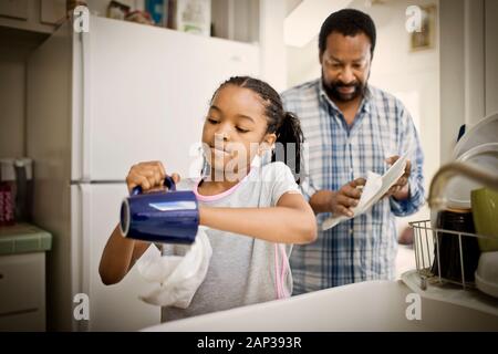 Ragazza che asciuga i piatti con il suo padre medio-adulto in una cucina. Foto Stock