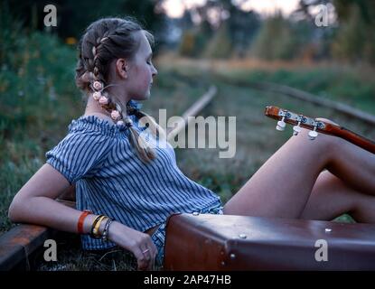 Una ragazza adolescente è seduto sui binari con una chitarra e una vecchia valigia Foto Stock