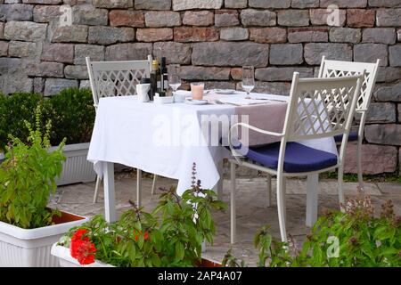 Tavolo con tovaglia bianca, posate e bicchieri da vino sulla veranda esterna estiva del ristorante. Muro di vecchia pietra testurizzata sullo sfondo. Foto Stock