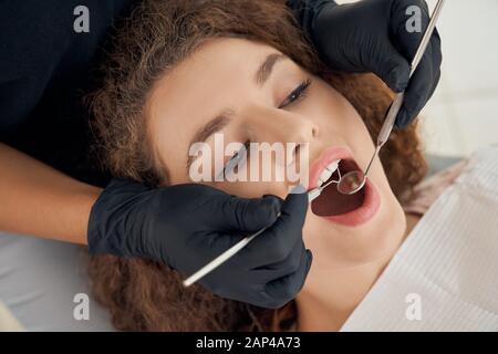 Dentista competente in guanti di gomma nera con specchio dentale e sonda che controlla i denti del paziente femminile. Ragazza affascinante con capelli ricci in regolare procedura di esame presso la clinica moderna. Foto Stock