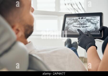 Giovane uomo con bearded seduto in sedia dentale e guardando la figura dei denti sulla compressa che tiene il dentista in guanti neri. Medico professionista che esamina la cavità orale del paziente maschio. Foto Stock