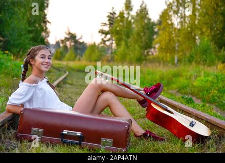 Una ragazza adolescente è seduto sui binari con una chitarra e una vecchia valigia Foto Stock