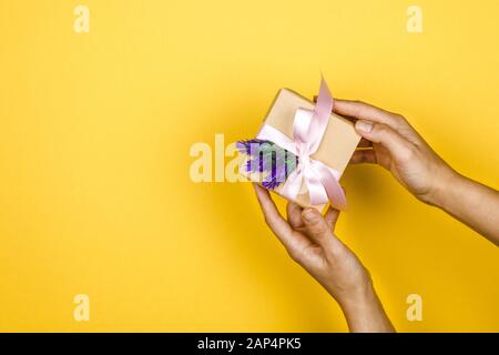 primo piano mani donna tenere una scatola regalo con un ramoscello di lavanda in un gesto di dare su sfondo giallo con spazio Foto Stock
