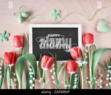 Composizione a molla con blackboard incorniciato con fiori di primavera, tulipani e il giglio della valle a pulire il tavolo di legno. Il testo 'Happy Holidays' sulla b Foto Stock