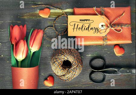 Dono avvolto con tag 'Happy Holidays', materiali di imballaggio, forbici, cavo a scacchi, cuori e tulipani rossi su una vecchia tavola in legno rustico. Concetto di pr Foto Stock
