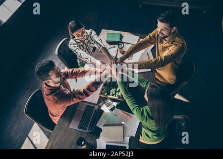 Vista dall'alto su alto angolo di quattro uomini d'affari allegri e professionisti che indossano informale abbigliamento da formalwear che mette insieme le palme sul desktop al moderno Foto Stock