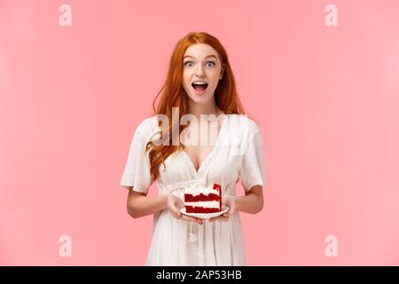 Sorpreso e divertito carismatico ragazza redhead spettegolare durante il party, tenendo la piastra con torta e bocca aperta affascinato come parlare su evento compleanno Foto Stock