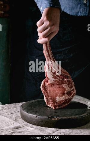 Uomo con le mani in mano azienda materie non cotti black angus tomahawk bistecca su ossa sulla tabella di lino inumidito. Stile rustico Foto Stock